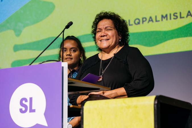 Aunty Jennifer Ketchell stands at a podium with a microphone. A young woman stands behind her.
