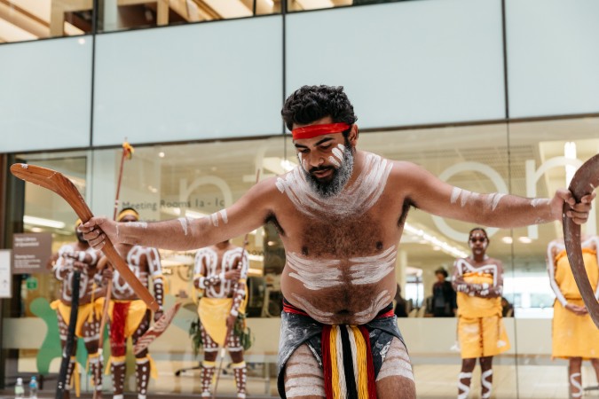 Bwgcolman Dancers perform in front of the glass walls of the State Library in full traditional dress.