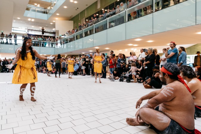 Bwgcolman and Nunukul Yuggera Dancers performing in the Knowledge Walk at the State Library. They are surrounded by a large audience watching from multiple levels of the library