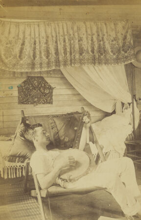 Sepia photograph depict John Henry Paul Berthon relaxing indoors in a reclining chair while playing a banjo.