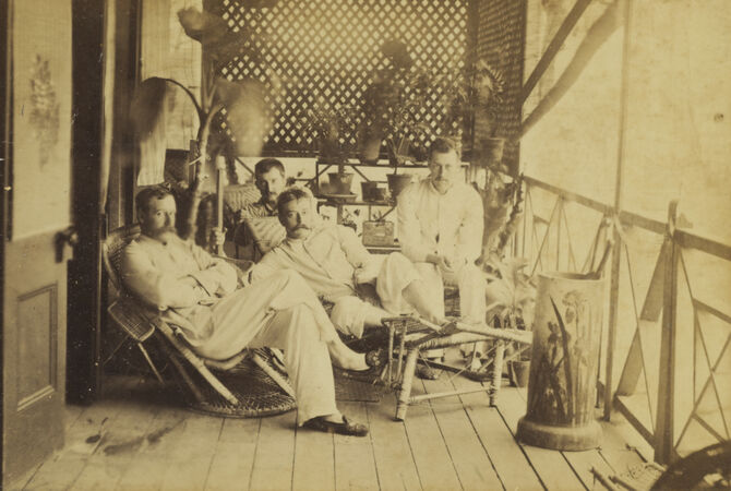 A sepia vintage photograph captures four European men relaxing on a shaded, plant-adorned tropical veranda, dressed in light-colored colonial leisure wear on a verandah in Cairns, Queensland, Australia.