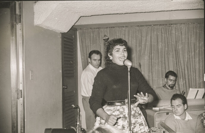 Black and white image of dark-haired woman singing at microphone with 3 musicians behind her