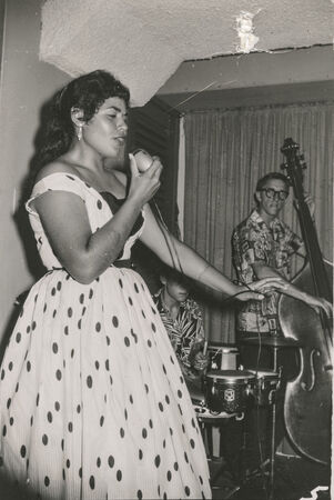 Black and white image of woman singing in spotted 1950s style dress and musicians playing behind her