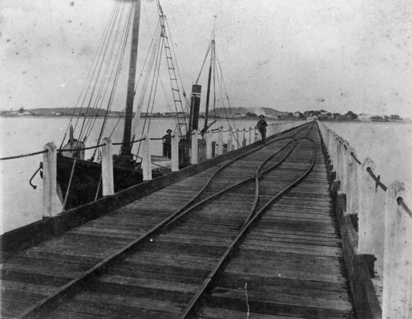 1800s view of jetty with small boat moored and railway tracks along the jetty
