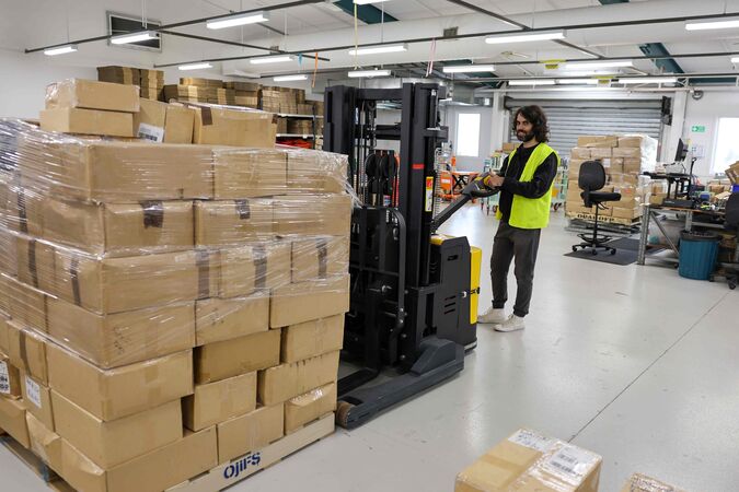 Man with dark hair and beard and hi-vis vest moving a pallet of boxes with a machine