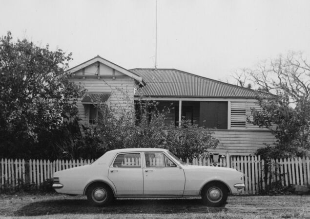 Black and white of single-storey weatherboard house with picket fence and car out front