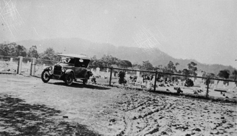 A vintage car parked beside a cemetery with mountains in the background.