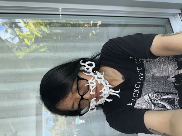 A photo of the artist Jent Do, who's face is visible, they are wearing black thick glasses and have long black hair. On their face is a interesting 3D printed wearable sculptures that looks like a beard.