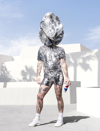  Photograph of a person wearing a sparkling shirt and shorts with a matching sparkling headdress that covers their face. They are holding a bejeweled cola cup.