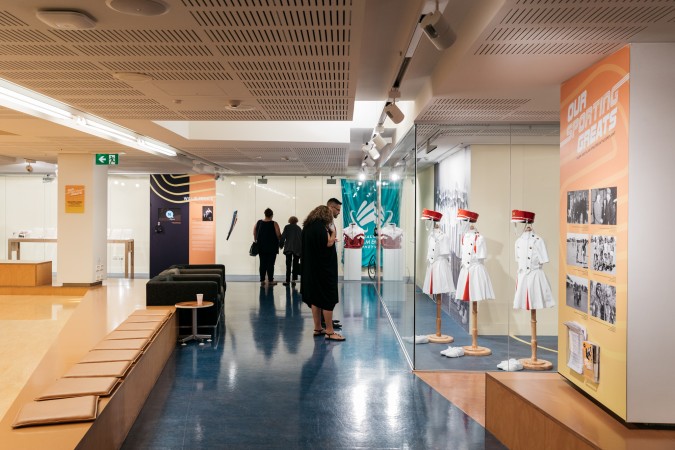 Exhibition space in kuril dhagun, a few people are walking through and looking at marching girl uniforms