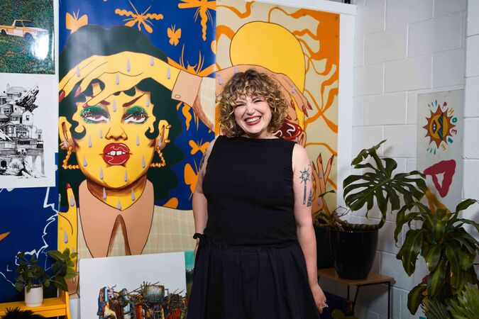 Curly-haired woman standing next to brightly coloured artwork of a woman holding a hand to her forehead