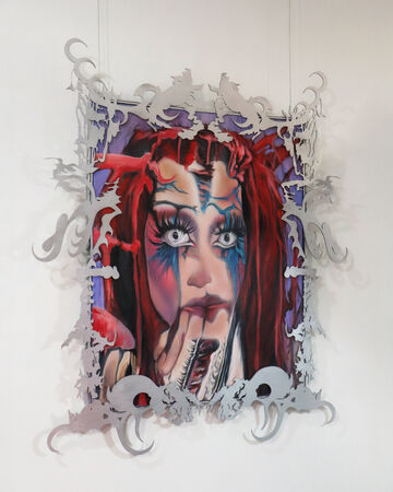  The image shows a striking portrait of a figure with intense, wide eyes and vivid red hair framed by intricate silver cutouts. The face features dramatic blue and red painted details, giving it a surreal and theatrical expression. This artwork is suspended against a plain background, with the ornate frame adding an otherworldly and decorative dimension.
