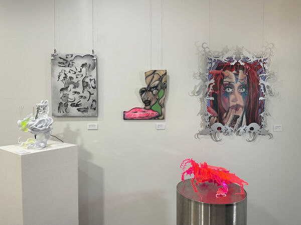  The photo shows a gallery display featuring four unique artworks. On the left is a sculptural piece with organic white forms surrounding a glowing yellow-green center. The wall displays include a monochrome abstract design, a portrait with distorted features and bright neon pink accents, and an intricate red-haired figure framed by ornate silver cutouts. To the right, a neon pink and red translucent sculpture sits atop a cylindrical pedestal, radiating energy and contrast.
