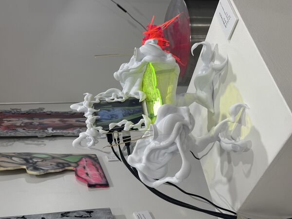 The artwork is a surreal sculpture with an organic, tentacle-like white frame surrounding a digital screen that displays a distorted face. It stands on claw-like legs and features a glowing yellow-green platform at its center, with thin sticks and bright red fibers adding striking details. The piece is displayed in a gallery setting, surrounded by colorful paintings on the walls.