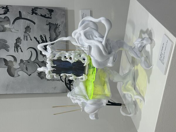 The artwork is a surreal sculpture with an organic, tentacle-like white frame surrounding a digital screen that displays a distorted face. It stands on claw-like legs and features a glowing yellow-green platform at its center, with thin sticks and bright red fibers adding striking details. The piece is displayed in a gallery setting, surrounded by colorful paintings on the walls.