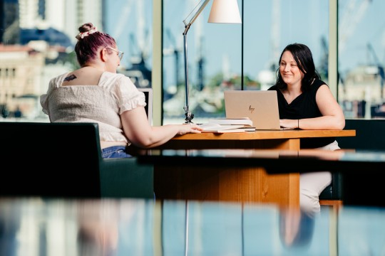 Two women sitting at a joint desk with a view of Brisbane city using laptops at State Library.