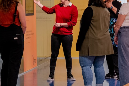 Showcase curator gestures to items behind glass in the Sovereign Stories showcase at State Library in front of a group of visitors.