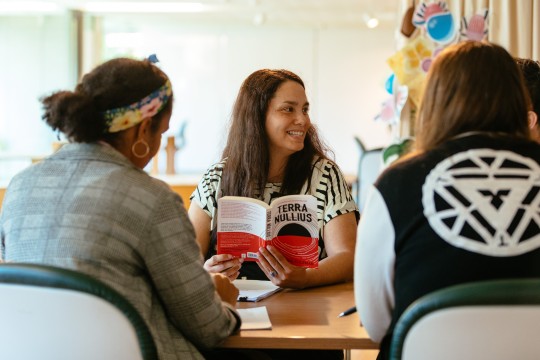 Three people are sitting at a table The one person facing the camera is holding the book Terra Nullius and is smiling to the side 