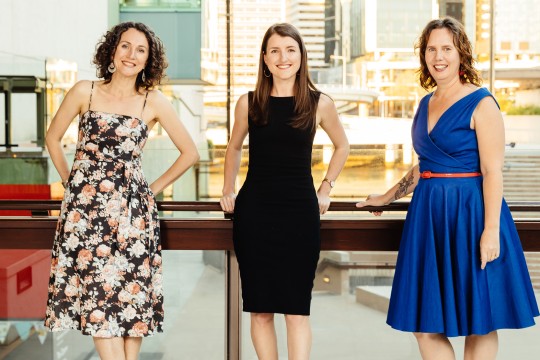 Kali Napier in a floral dress, Ella Jeffery in black and Tabitha Bird in blue stand smiling at the railing of the Queensland Terrace