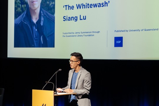 Siang Lu stands onstage behind a lectern with a blue and yellow pineapple on the front Behind him is the name of his winning novel onscreen The Whitewash