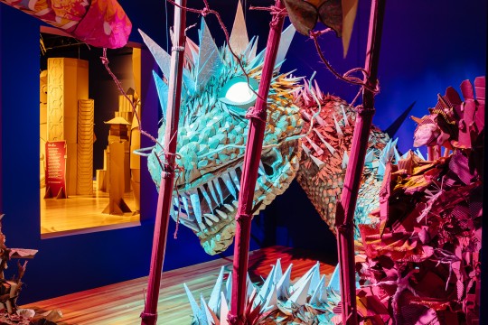 A large cardboard dragon seen through cardboard bamboo trunks with cardboard koi fish floating in the air.