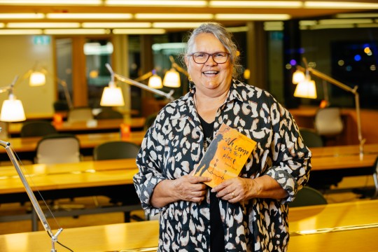 Debra Dank at the 2023 Queensland Literary Awards. She stands in a study room with lamps. She is smiling and holding a copy of her book.