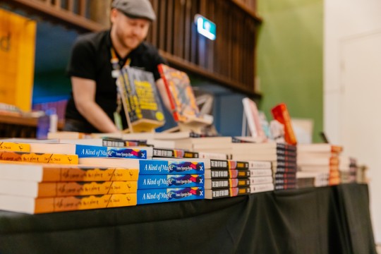 A person stands behind a table with piles of books on it at a pop-up books shop.