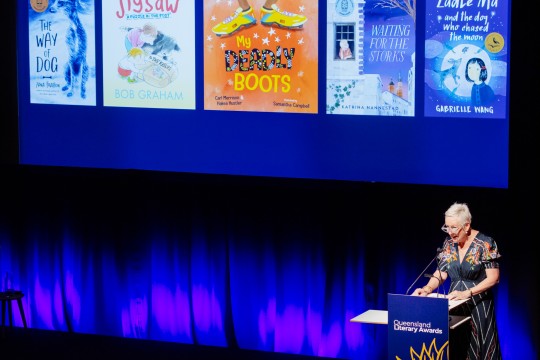 Susan Hocking stands onstage with an auditorium screen behind her showing 5 kids' books