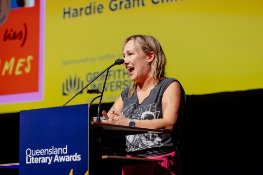 Biffy James stands at a lectern. She wears a grey top and her book is on a screen behind her.