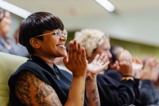 A woman with short hair and glasses sits in an auditorium smiling and applauding
