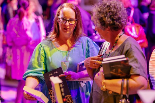 Two people carrying books and browsing a book stall at a night-time event