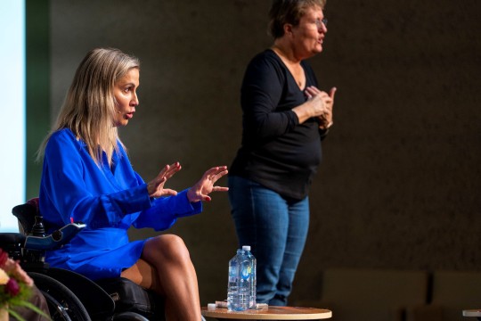 Karni Liddell wears an electric blue dress and talks to the audience. An Auslan interpreter is in the background