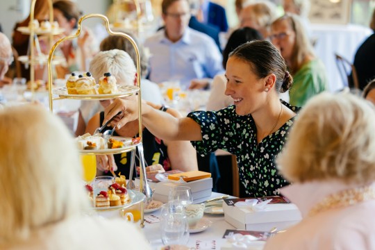 Person using tongs to pick up a small cake from a high tea tray.