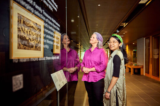 Two women looking at artworks hanging on a wall behind glass.