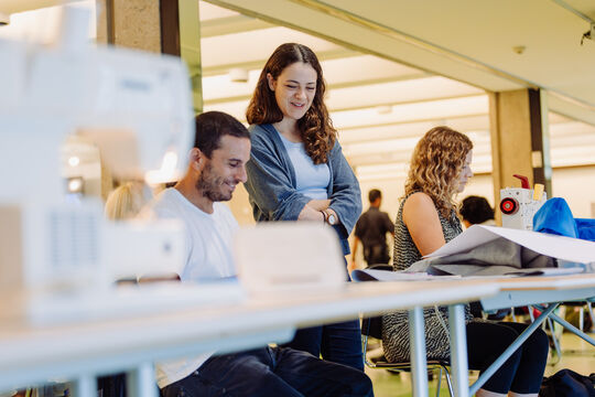 Two people smile and discuss sewing, while another person sits next to them concentrating at a sewing machine