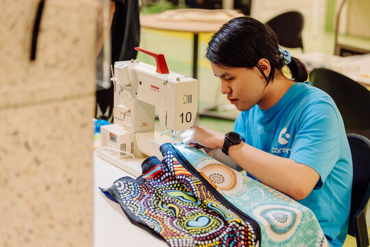 A person who is wearing a ligth blue shirt, and has dark hair tied back and is sewing at a table. A sewing machine is visible and a colourful piece of fabric. 