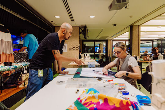 People sewing at The Edge during Threads 2024, someone is visible on the right hand side using a ruler to cut out a pattern, someone on the left hand side is standing over the table and pointing at the pattern to help.