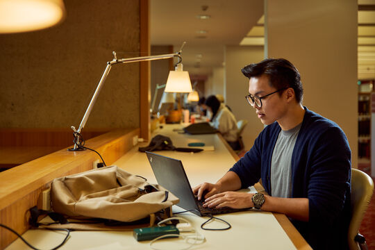 A person working on a laptop at a long desk in a quiet study area, with a backpack and lamp nearby.
