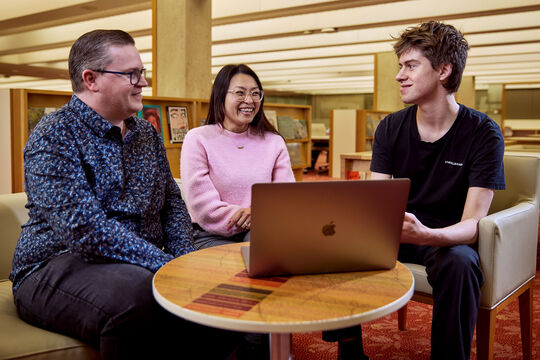 Three people sitting together in the State Library with a laptop open in front of them.