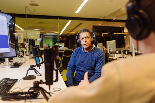 Man in a blue sweater wearing headphones is interviewed by a man on the far right of the frame. They use podcasting microphones