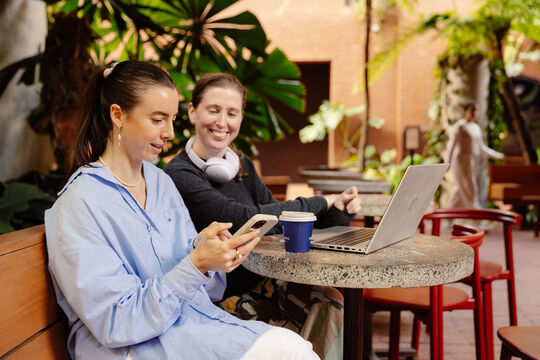 Two women at a cafe, one with a laptop and the other with a mobile phone.