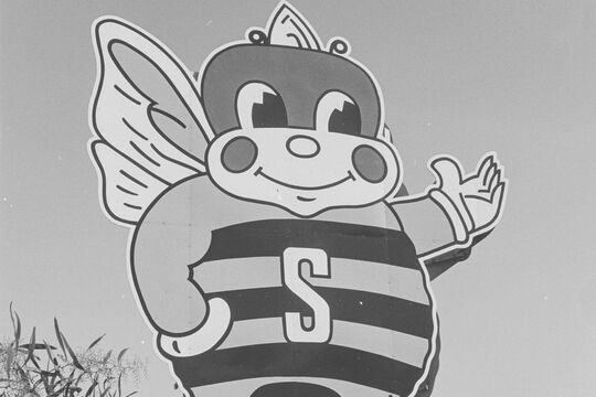 Black and white photograph of 3 people waving in front of a large bee sign.