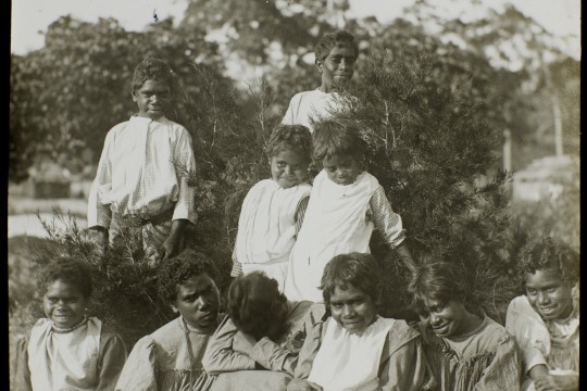 Group of First Nations children in front of shrubs, four children are standing with six children sitting on the ground in front.