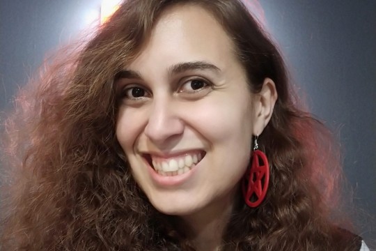 A photograph of writer Allanah Hunt who has wavy brown hair haloed by soft lighting and wears red pentagram earrings