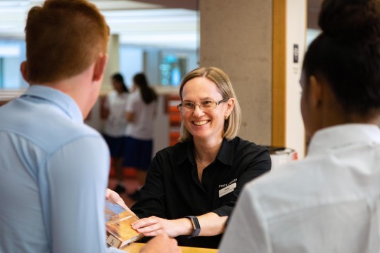 State Library staff member smiling and passing two visitors a book from behind a counter. 