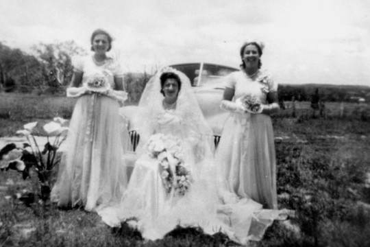 Bride and bridesmaids standing in front of a Holden, ca. 1951. The bride is sat between her two standing bridesmaids wearing a big white bridal gown, holding a bouquet.