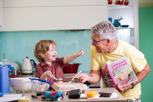 Young child and older man smiling and cooking in the kitchen. The man is holding a cupcake book book while the young child holds a spoon and points. 