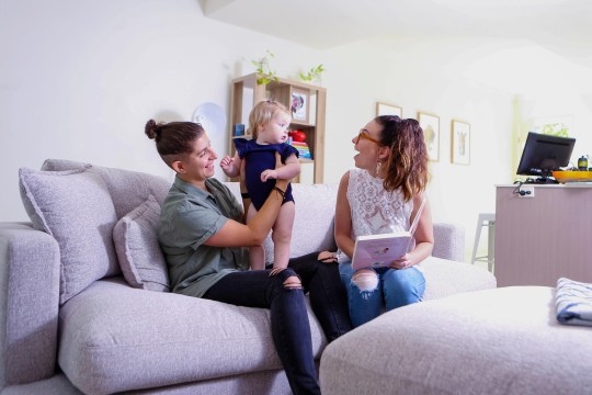 Two people sitting on a couch at home with a book One person has a young child on her knee