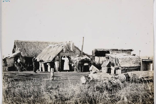 Photo of people standing outside buildings with thatch roof and lean-to buildings attached