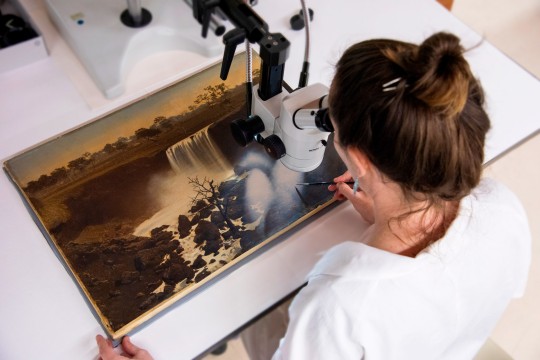 Over the shoulder shot of a State Library staff member looking at a painting through a microscope and painting small details with a paintbrush.